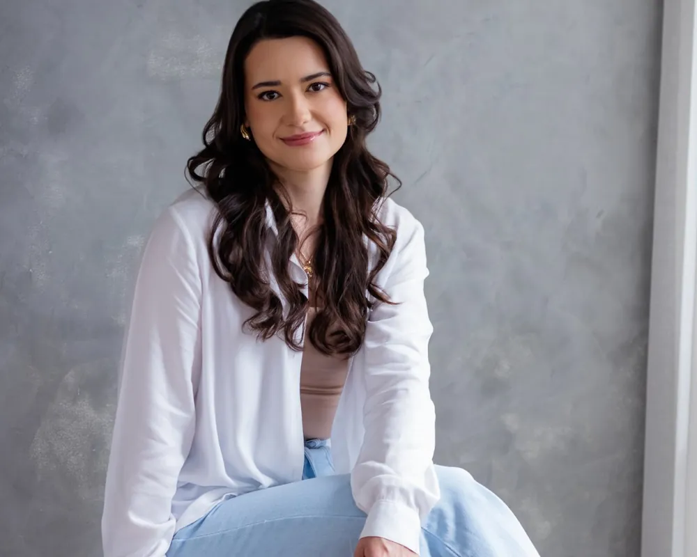 Isabela Matoso sentada em um banco alto, de camisa branca e calça jeans